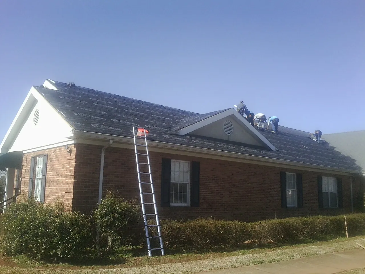 Crew tearing shingles off a brick ranch roof with a ladder set — Emergency Roof Repair work by our crew in Mountain Rest