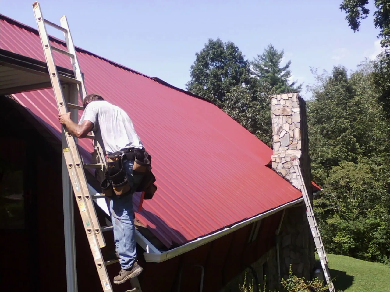 Roofer on a ladder beside a red metal roof and stone chimney — Soffit & Fascia Repair by Roofing Brothers in Mountain Rest